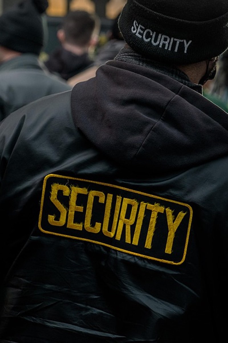 A guard in a marked black security jacket and beanie, working a crowd at an outdoor event.
