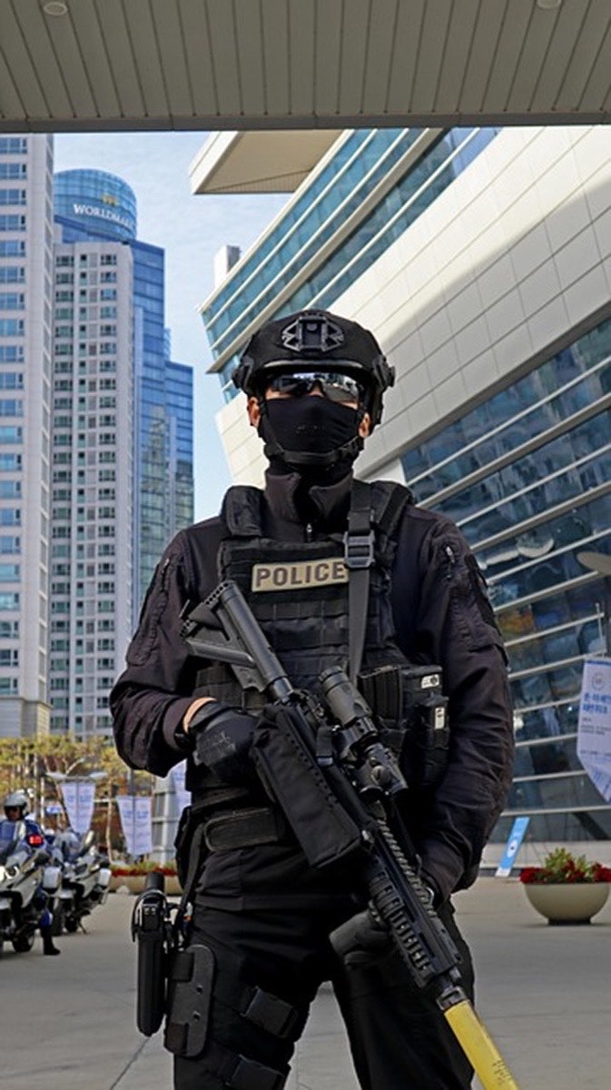An officer in tactical body armour and helmet holding a rifle outside a commercial building.